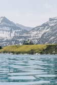 A large building resembling a lodge or hotel is situated on a grassy hill overlooking a calm, expansive body of water. Snow-capped mountains rise in the background under a pale blue sky.