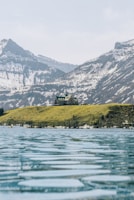 A large building resembling a lodge or hotel is situated on a grassy hill overlooking a calm, expansive body of water. Snow-capped mountains rise in the background under a pale blue sky.
