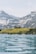 A large building resembling a lodge or hotel is situated on a grassy hill overlooking a calm, expansive body of water. Snow-capped mountains rise in the background under a pale blue sky.