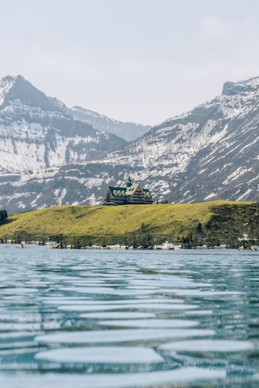 A large building resembling a lodge or hotel is situated on a grassy hill overlooking a calm, expansive body of water. Snow-capped mountains rise in the background under a pale blue sky.