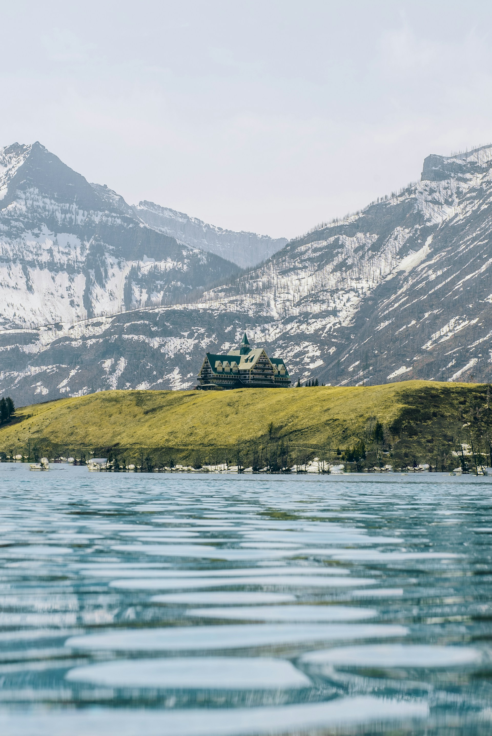 A large building resembling a lodge or hotel is situated on a grassy hill overlooking a calm, expansive body of water. Snow-capped mountains rise in the background under a pale blue sky.