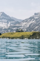 A large building resembling a lodge or hotel is situated on a grassy hill overlooking a calm, expansive body of water. Snow-capped mountains rise in the background under a pale blue sky.