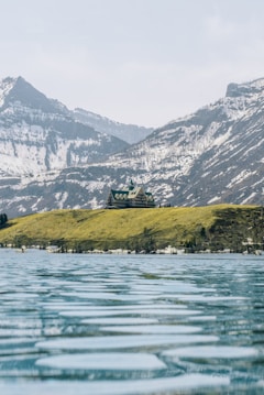 A large building resembling a lodge or hotel is situated on a grassy hill overlooking a calm, expansive body of water. Snow-capped mountains rise in the background under a pale blue sky.