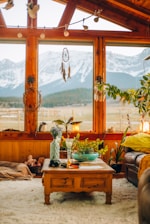 A cozy living room with large wooden-framed windows offering a view of snow-capped mountains. The room is decorated with various plants, a dreamcatcher, and a comfortable seating area. A wooden coffee table in the center holds a vase with flowers, a green bowl, and other small items. The warm earthy tones and soft textures create a welcoming atmosphere.