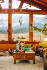 A cozy living room with large wooden-framed windows offering a view of snow-capped mountains. The room is decorated with various plants, a dreamcatcher, and a comfortable seating area. A wooden coffee table in the center holds a vase with flowers, a green bowl, and other small items. The warm earthy tones and soft textures create a welcoming atmosphere.