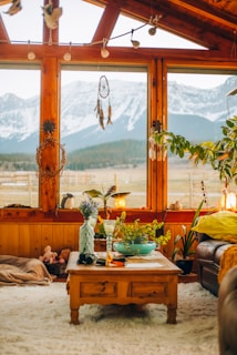 A cozy living room with large wooden-framed windows offering a view of snow-capped mountains. The room is decorated with various plants, a dreamcatcher, and a comfortable seating area. A wooden coffee table in the center holds a vase with flowers, a green bowl, and other small items. The warm earthy tones and soft textures create a welcoming atmosphere.