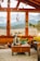A cozy living room with large wooden-framed windows offering a view of snow-capped mountains. The room is decorated with various plants, a dreamcatcher, and a comfortable seating area. A wooden coffee table in the center holds a vase with flowers, a green bowl, and other small items. The warm earthy tones and soft textures create a welcoming atmosphere.