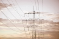 A series of tall electrical power lines stretch across the sky, supported by metal lattice towers. The landscape has a serene quality with soft, cloudy skies in the background, creating a contrast between nature and industry.
