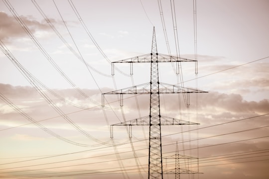 A series of tall electrical power lines stretch across the sky, supported by metal lattice towers. The landscape has a serene quality with soft, cloudy skies in the background, creating a contrast between nature and industry.