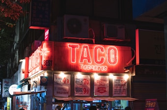 A night scene of a street food stall with a prominent neon sign that reads 'TACO' and 'TOBACCOS.' The stall advertises food items like 'OLDIES TACO' and 'TACO RICE' with prices. There's a note saying 'Sorry we're SOLD OUT.' The environment includes other lighted signage and a visible air conditioning unit above.