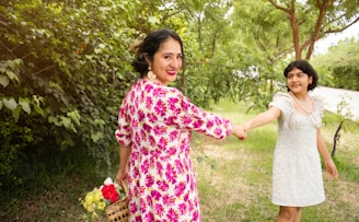A mother and daughter holding hands in a sun-dappled garden filled with pink and gold flowers.