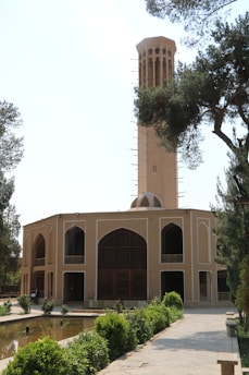 A tall wind tower rises above a traditional Middle Eastern building, surrounded by lush greenery and a reflective water feature. The structure features arched doorways and elaborate lattice work, characteristic of regional architecture.