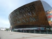 A large, modern architectural structure with a curved facade made of metallic, copper-toned panels. The building has large lettering cut out in two languages across the exterior. The surrounding area is a spacious open plaza with paving, and a futuristic, undulating white structure is visible in the background.