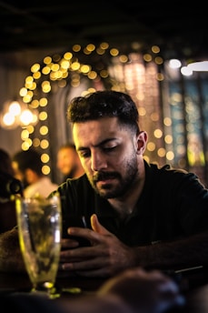 A man with a beard is focused on his smartphone at a dimly lit bar or restaurant. The background features out-of-focus warm fairy lights and ambient lighting creating a cozy atmosphere.