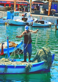 A fisherman stands on a small boat in a harbor, handling fishing nets. The boat is blue and white, with additional nets piled up at the back. Several other boats are docked nearby, with buildings and people visible in the background. The water reflects the vibrant colors of the scene.