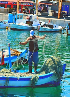 A fisherman stands on a small boat in a harbor, handling fishing nets. The boat is blue and white, with additional nets piled up at the back. Several other boats are docked nearby, with buildings and people visible in the background. The water reflects the vibrant colors of the scene.