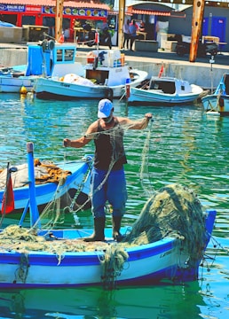 A fisherman stands on a small boat in a harbor, handling fishing nets. The boat is blue and white, with additional nets piled up at the back. Several other boats are docked nearby, with buildings and people visible in the background. The water reflects the vibrant colors of the scene.