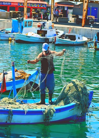A fisherman stands on a small boat in a harbor, handling fishing nets. The boat is blue and white, with additional nets piled up at the back. Several other boats are docked nearby, with buildings and people visible in the background. The water reflects the vibrant colors of the scene.