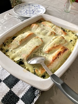 A white baking dish containing cooked salmon fillets in a creamy sauce, placed on a dining table. The salmon is slightly browned and layered over spinach. A silver serving spoon rests on the dish, and a black and white striped kitchen towel lies beside it. In the background, a blue and white patterned plate sits on the table next to a small glass vase with flowers.