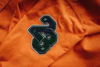 A close-up view of an orange corduroy fabric with a large black and green embroidered patch featuring the number 3. The texture of the fabric is visible, and the patch is prominently centered.