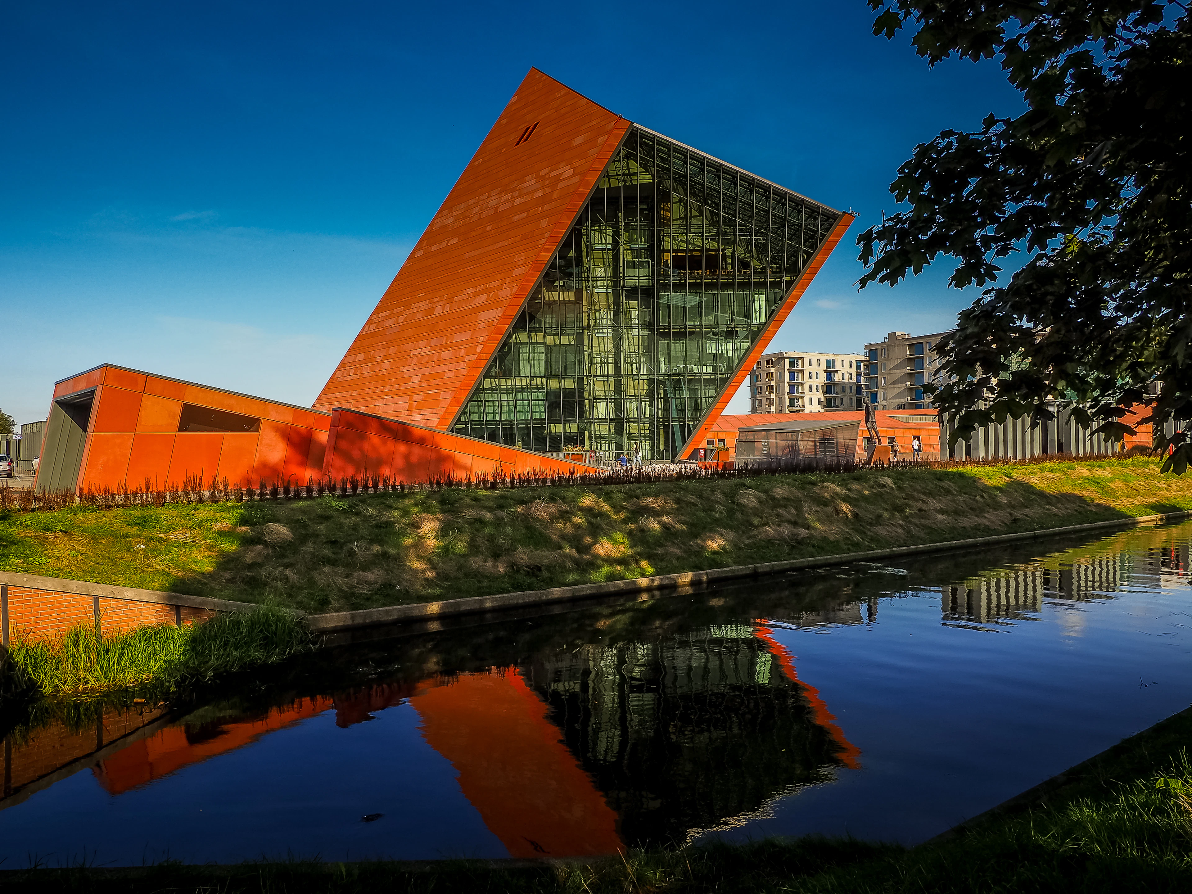 The Museum of the Second World War in Gdansk, its rust-red wedge leaning back over a wall of glass and doubled in the still canal below, with the low red plaza wall and a grass bank running along the waterside under a hard blue sky.