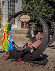A person dressed in colorful, traditional clothing sits on a modern, circular metal bench in an urban setting. A vibrant flag with a distinctive pattern is draped over the bench. The background features a building with large windows, a sign that reads 'Jarmark Sw. Dominika,' and a mannequin dressed in folk costume.