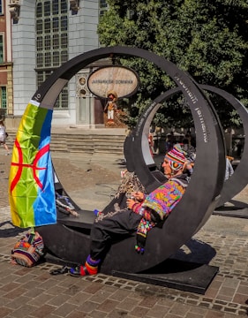 A person dressed in colorful, traditional clothing sits on a modern, circular metal bench in an urban setting. A vibrant flag with a distinctive pattern is draped over the bench. The background features a building with large windows, a sign that reads 'Jarmark Sw. Dominika,' and a mannequin dressed in folk costume.
