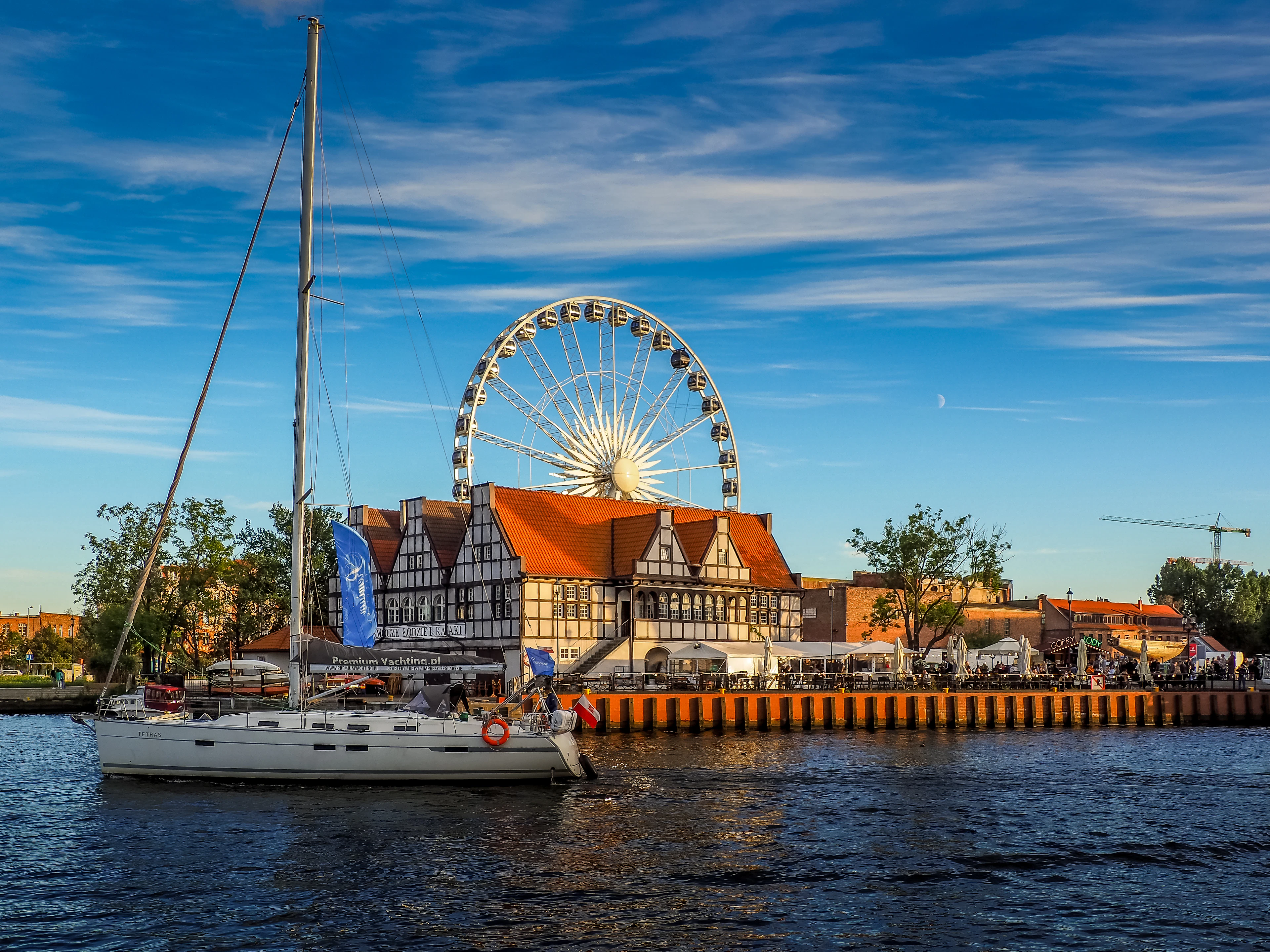 A yacht motoring down the Motlawa in Gdansk past a half-timbered waterfront building with its terrace tables full, the white AmberSky ferris wheel turning behind it in late golden light.