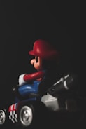 A toy figure is seated in a small racing kart, predominantly featuring red and blue colors with a black and white checkered pattern on the side. The figure is wearing a red hat and blue overalls.