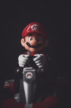 A figure with a red cap featuring an 'M' emblem, a large mustache, and a red and blue outfit is sitting in a kart. The character is holding the steering wheel, and the image is set against a dark background, highlighting the figure.