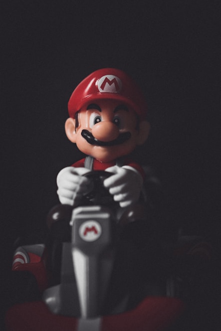 A figure with a red cap featuring an 'M' emblem, a large mustache, and a red and blue outfit is sitting in a kart. The character is holding the steering wheel, and the image is set against a dark background, highlighting the figure.