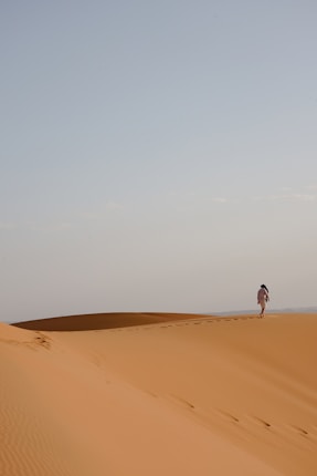 A solitary figure walks along the crest of a vast sand dune, leaving footsteps behind. The expansive desert landscape stretches under a clear, light blue sky.
