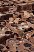 A traditional tannery with numerous circular stone vats filled with various colored liquids arranged in a geometric pattern. Two workers, one standing wearing a straw hat and rubber overalls, and one seated near the vats, appear to be engaged in the process of treating hides. Large pieces of leather are draped over some of the vats, drying under the sun, amidst the earthy tones of the environment.