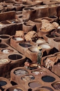 A traditional tannery with numerous circular stone vats filled with various colored liquids arranged in a geometric pattern. Two workers, one standing wearing a straw hat and rubber overalls, and one seated near the vats, appear to be engaged in the process of treating hides. Large pieces of leather are draped over some of the vats, drying under the sun, amidst the earthy tones of the environment.