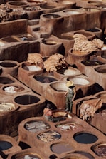 A traditional tannery with numerous circular stone vats filled with various colored liquids arranged in a geometric pattern. Two workers, one standing wearing a straw hat and rubber overalls, and one seated near the vats, appear to be engaged in the process of treating hides. Large pieces of leather are draped over some of the vats, drying under the sun, amidst the earthy tones of the environment.