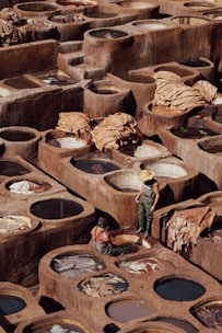 A traditional tannery with numerous circular stone vats filled with various colored liquids arranged in a geometric pattern. Two workers, one standing wearing a straw hat and rubber overalls, and one seated near the vats, appear to be engaged in the process of treating hides. Large pieces of leather are draped over some of the vats, drying under the sun, amidst the earthy tones of the environment.