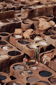 A traditional tannery with numerous circular stone vats filled with various colored liquids arranged in a geometric pattern. Two workers, one standing wearing a straw hat and rubber overalls, and one seated near the vats, appear to be engaged in the process of treating hides. Large pieces of leather are draped over some of the vats, drying under the sun, amidst the earthy tones of the environment.
