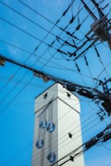 A tall, light-colored building adorned with a mural of large water droplets on its side. Numerous overhead electrical wires and power lines crisscross the foreground against a clear blue sky.