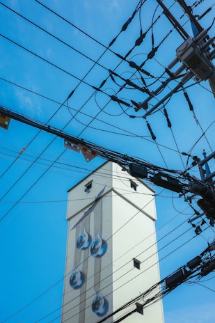 A tall, light-colored building adorned with a mural of large water droplets on its side. Numerous overhead electrical wires and power lines crisscross the foreground against a clear blue sky.