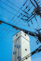 A tall, light-colored building adorned with a mural of large water droplets on its side. Numerous overhead electrical wires and power lines crisscross the foreground against a clear blue sky.
