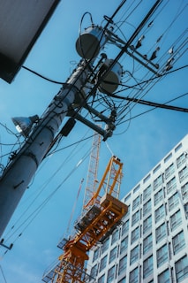 A large yellow construction crane stands next to a high-rise building under a clear blue sky. Numerous wires and a utility pole are prominently featured, intersecting in the foreground. The building is modern with a grid of windows, and the crane is partially positioned as if carrying something.