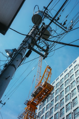 A large yellow construction crane stands next to a high-rise building under a clear blue sky. Numerous wires and a utility pole are prominently featured, intersecting in the foreground. The building is modern with a grid of windows, and the crane is partially positioned as if carrying something.