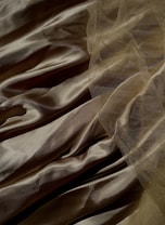 Silky fabric in a golden hue is draped and layered with soft, gentle folds. The surface has a subtle sheen, reflecting light across its smooth texture. A delicate mesh overlay adds depth, creating intricate patterns and enhancing the visual interest.