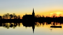 A tranquil silhouette of a church and trees is reflected in a calm body of water during a sunset. The sky is a gradient of yellow and orange hues, casting warm tones across the scene.