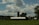 A rural landscape featuring a farm with a large barn and two silos set against a backdrop of rolling hills. The sky is filled with fluffy clouds, and the foreground shows a lush green field with crops. Trees are scattered throughout the scene, adding a natural element.