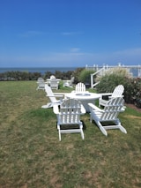 A scenic outdoor setting featuring several white Adirondack chairs arranged around a round table on a grassy lawn. The background includes lush greenery and a wooden fence, with a clear view of the ocean under a bright blue sky. The area is well-maintained, suggesting a peaceful and inviting atmosphere.