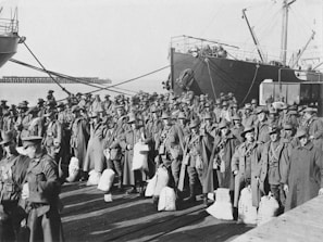 A large group of soldiers in uniform and wide-brimmed hats are standing in rows on a dock. They are carrying bags, and there are two large ships docked nearby. The background shows the open water and part of a pier extending into the distance.