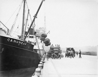 A dockside scene featuring a ship named CANONBAR moored alongside a quay. Workers are engaged in unloading large bundles from the ship onto trucks waiting on the dock. The trucks have visible company logos, and several workers are moving about or supervising the operations.
