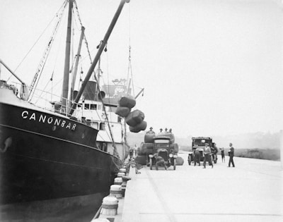 A dockside scene featuring a ship named CANONBAR moored alongside a quay. Workers are engaged in unloading large bundles from the ship onto trucks waiting on the dock. The trucks have visible company logos, and several workers are moving about or supervising the operations.