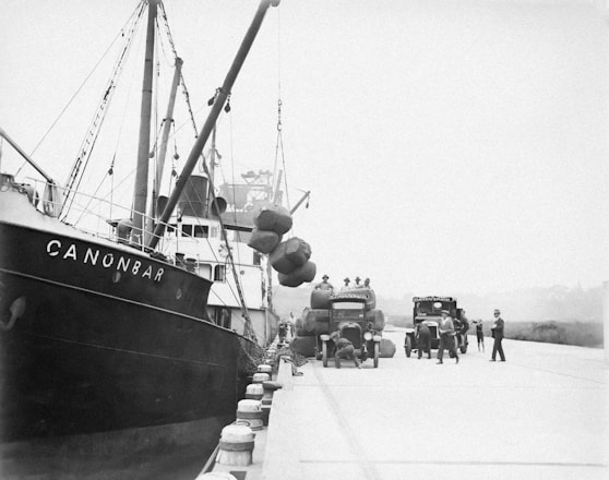 A dockside scene featuring a ship named CANONBAR moored alongside a quay. Workers are engaged in unloading large bundles from the ship onto trucks waiting on the dock. The trucks have visible company logos, and several workers are moving about or supervising the operations.